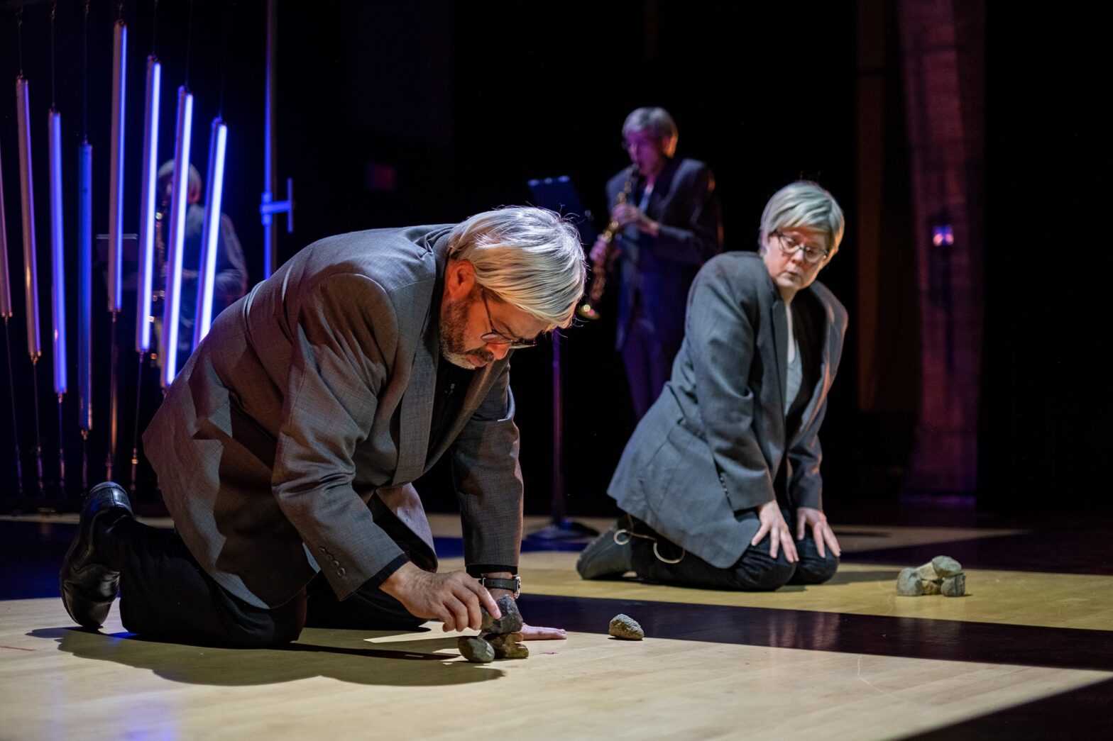 closeup of Arturo O'Farrill and Tony Arnold onstage in suits and wigs kneeling and stacking rocks